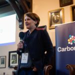 Nicky holds a microphone and talks to the crowd at the venture builder's showcase day. in the background, the carbon13 logo is visible on a banner in the Cambridge Union