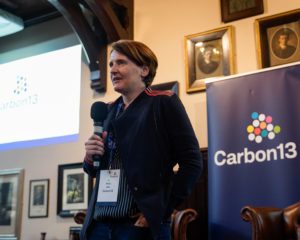 Nicky holds a microphone and talks to the crowd at the venture builder's showcase day. in the background, the carbon13 logo is visible on a banner in the Cambridge Union